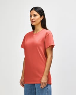 A model wearing a coral Gildan Heavy Cotton Adult T-Shirt, paired with light blue jeans. The shirt has a logo.