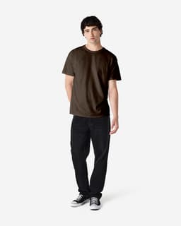 A man stands wearing a dark brown Gildan Heavy Cotton adult t-shirt and black jeans, with a logo visible on the shirt.