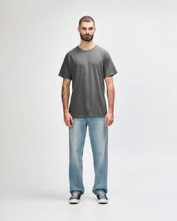 A grey adult t-shirt displayed on a male model, paired with black trousers and sneakers. The shirt has a logo.