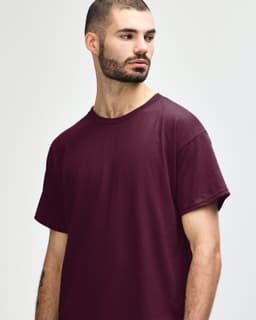 A maroon Gildan Heavy Cotton Adult T-Shirt displayed on a model with short, curly hair. The shirt has a logo.
