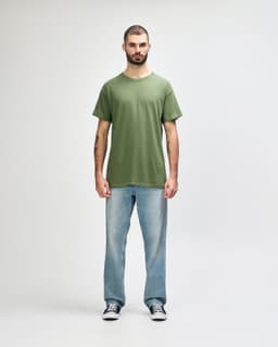 A green Gildan Heavy Cotton Adult T-Shirt, worn by a model paired with black jeans and sneakers, standing against a white background.