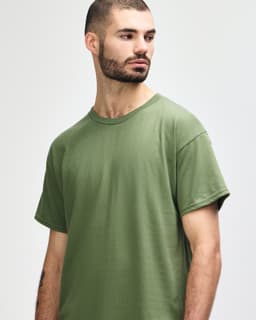 A man models a Gildan Heavy Cotton Adult T-Shirt in olive green, featuring a simple design and a logo.