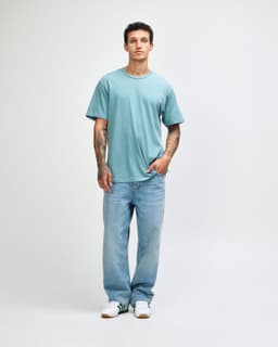 A man wearing a light blue unisex short sleeve t-shirt with denim jeans, standing against a plain background.