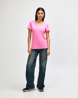 A woman stands wearing a pink Gildan Softstyle ladies' V-neck T-shirt and blue jeans, with white sneakers.