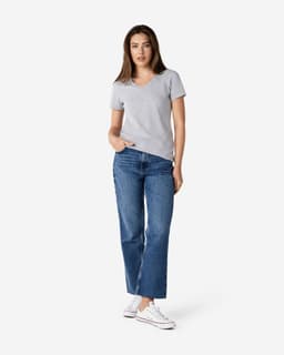 A woman stands wearing a grey Gildan Softstyle ladies' v-neck t-shirt and blue jeans, paired with white sneakers.
