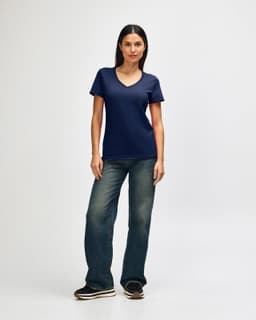 A woman is wearing a navy Gildan Softstyle ladies' V-neck T-shirt, paired with blue jeans and white sneakers.