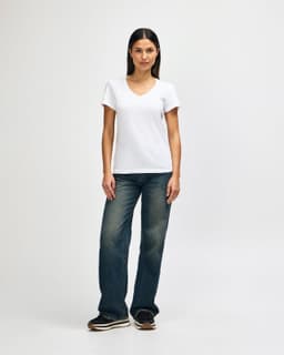 A model wearing a white Gildan Softstyle Ladies' V-Neck T-Shirt paired with blue jeans and white sneakers.