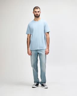 A midweight adult T-shirt in light blue, worn by a model with dark pants and sneakers, featuring a logo.