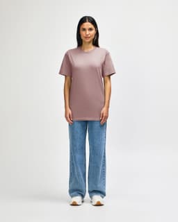 A midweight adult T-shirt in soft pink, worn with black jeans and sneakers. Features a logo.