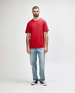 A red Gildan Softstyle adult midweight T-shirt displayed on a model wearing black jeans and shoes. The shirt has a logo.