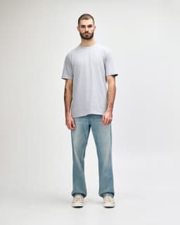 A man stands wearing a light grey Gildan Softstyle adult midweight T-shirt and blue jeans. The shirt has a logo.