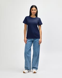 A woman wearing a navy Gildan Softstyle ladies midweight T-shirt, paired with blue jeans and white sneakers.