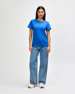 A woman stands wearing a blue Gildan Softstyle ladies midweight T-shirt and jeans, paired with white sneakers.