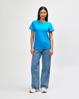 A woman stands wearing a teal Gildan Softstyle Ladies Midweight T-Shirt paired with blue jeans and white sneakers.