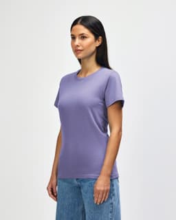 A woman wearing a purple Gildan Softstyle Ladies Midweight T-Shirt paired with blue jeans, smiling at the camera.