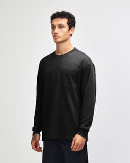 A black long-sleeve t-shirt featuring a pocket, modelled by a person with curly hair. The shirt has a logo.