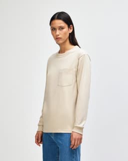 A long-sleeve beige t-shirt with a pocket and a crew neck, displayed by a model wearing olive green pants.