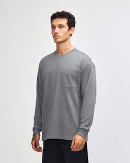 A man is wearing a grey long-sleeve t-shirt with a pocket, standing in a neutral backdrop. The shirt has a logo.