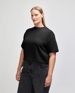 A black mid-length boxy top displayed on a model, showing a side view with short sleeves. The top has a logo.