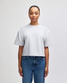 A woman stands wearing a grey mid-length boxy top, paired with light blue jeans. The top features a logo.