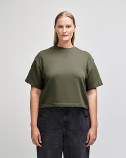 A woman wearing an olive green mid-length boxy top with short sleeves, paired with light blue jeans. The top features a logo.