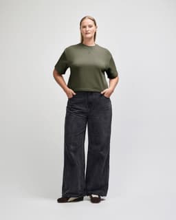 A woman stands wearing an olive green boxy top and light blue wide-leg jeans. The outfit is casual and stylish.