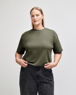 A women's mid-length boxy top in dark green, featuring a relaxed fit and short sleeves. The model is standing confidently.