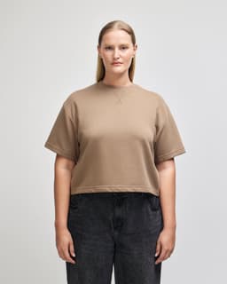 A mid-length boxy top in a tan colour, featuring short sleeves and a relaxed fit. The item has a logo.