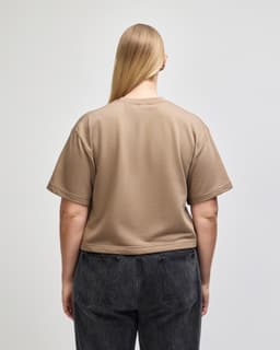 A back view of a beige mid-length boxy top with short sleeves, paired with dark grey pants. The top features a logo.