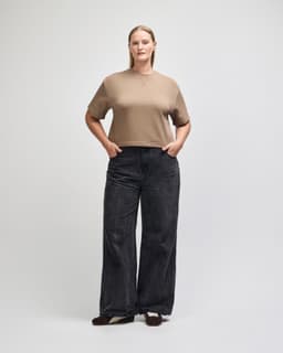 A mid-length boxy top in beige, worn with black wide-legged jeans. The top has a brand logo on it.