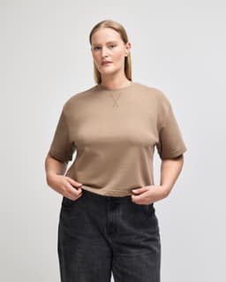 A mid-length boxy top in a light brown colour, featuring short sleeves and a round neckline. The model wears dark jeans.