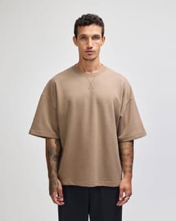 A unisex oversized boxy top in a tan colour, featuring a casual fit and short sleeves. It has a visible logo.