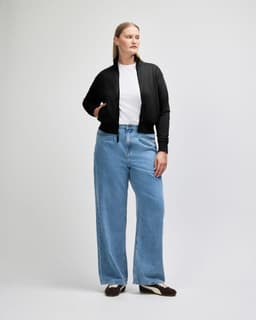 A woman wearing a black full-zip jacket and blue jeans, standing with one hand on her hip. The jacket has a logo.