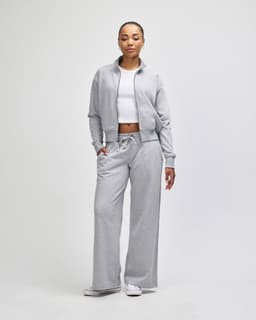 A woman stands wearing a grey full-zip jacket and matching wide-leg pants over a white top. The outfit has a logo.