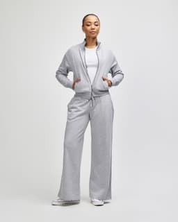 A woman stands in a light grey full-zip jacket paired with matching wide-leg pants. The jacket has a logo.