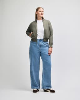 A woman wearing an olive green full-zip jacket over a white t-shirt, paired with light blue wide-leg jeans.