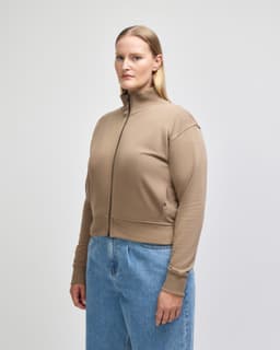 A women's full-zip jacket in beige, featuring a high collar and a fitted silhouette, worn with blue jeans.