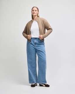 A woman wears a beige full-zip jacket over a white top, paired with wide-legged blue jeans. The jacket features a logo.
