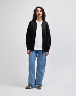 A black unisex bomber jacket is worn by a model, paired with light blue jeans and white shoes. The jacket has a logo.