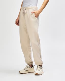 Unisex sweatpants in a light beige colour, featuring a logo, styled with a pink top and white sneakers.