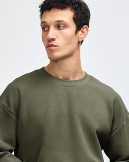A man wearing an olive green unisex crewneck sweatshirt. The sweatshirt features a loose fit and has a logo.