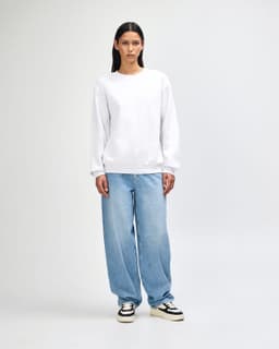 A unisex crewneck sweatshirt in white, paired with light blue baggy jeans, worn by a model. Features a branding logo.