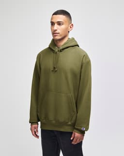 A green hooded sweatshirt with a front pocket and a logo on the sleeve, worn by a model standing against a plain background.