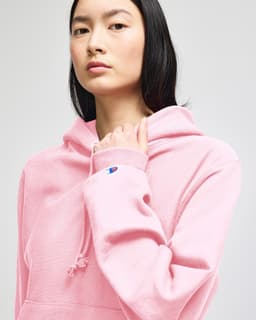 A pink hoodie with a drawstring hood and a logo on the sleeve. The model is looking directly at the camera.