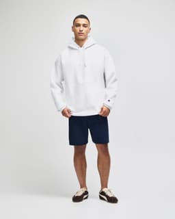 A white pullover hooded sweatshirt paired with navy shorts, featured on a model. The sweatshirt has a logo.