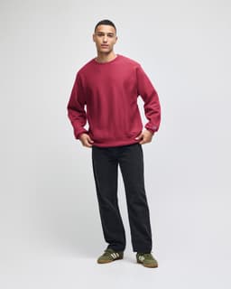 A red crew sweatshirt is worn by a model standing in a casual pose, paired with dark trousers and green shoes.