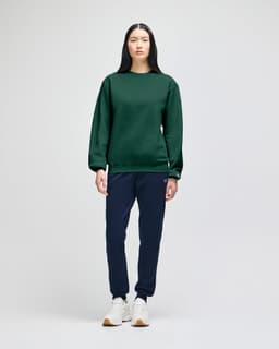 An olive green crewneck sweatshirt is worn with blue jeans, featuring a logo on the sleeve.