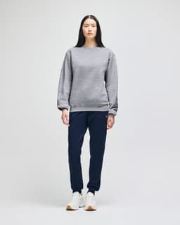 A grey crewneck sweatshirt worn by a model, paired with black pants and shoes, featuring a logo on the garment.