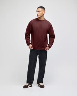 A model wearing a burgundy crewneck sweatshirt and dark pants, featuring a logo on the sleeve.