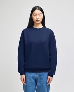 A navy adult crewneck sweatshirt is worn by a model, featuring a logo on the sleeve. The model pairs it with denim jeans.